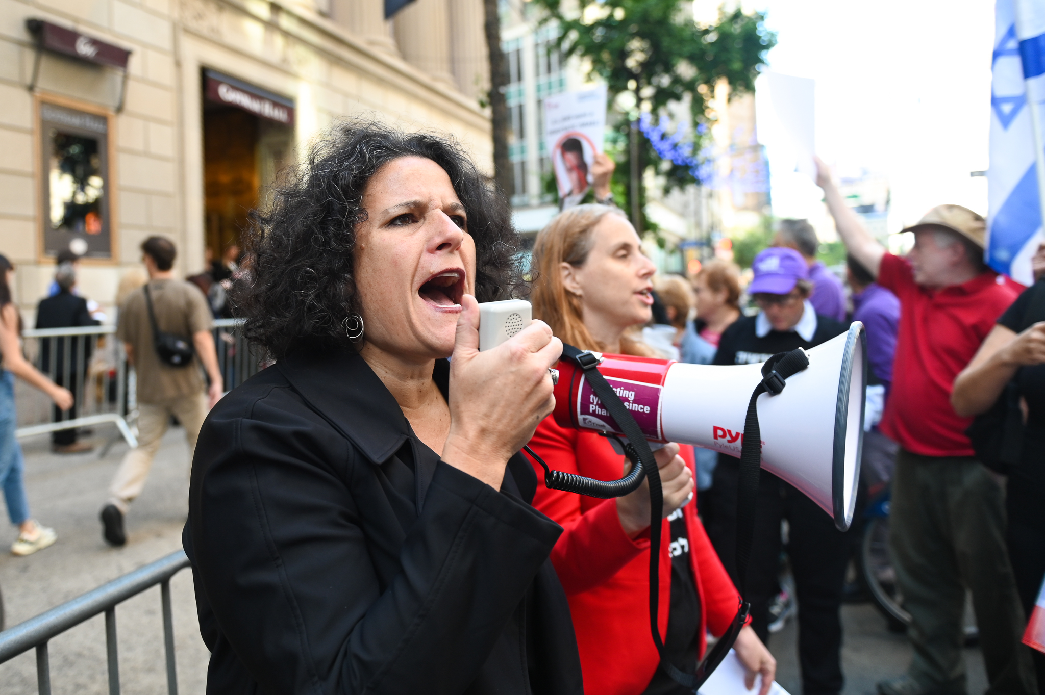 A woman in kippah and blazer speaks into a megaphone with protest in background